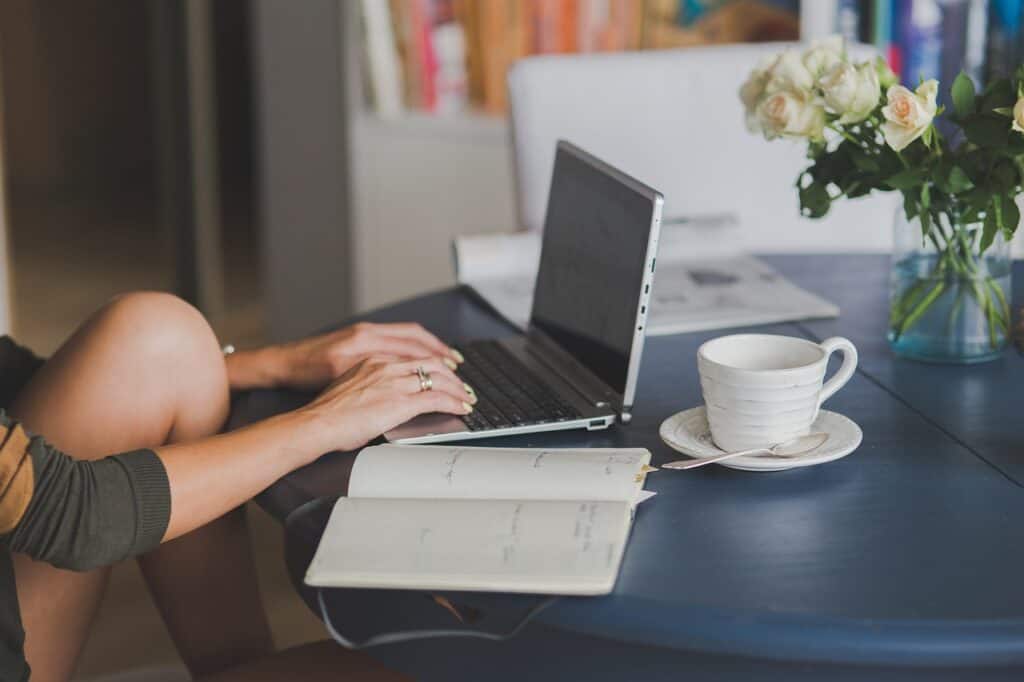 Person typing on a laptop at a table with a notebook and cup of coffee nearby.